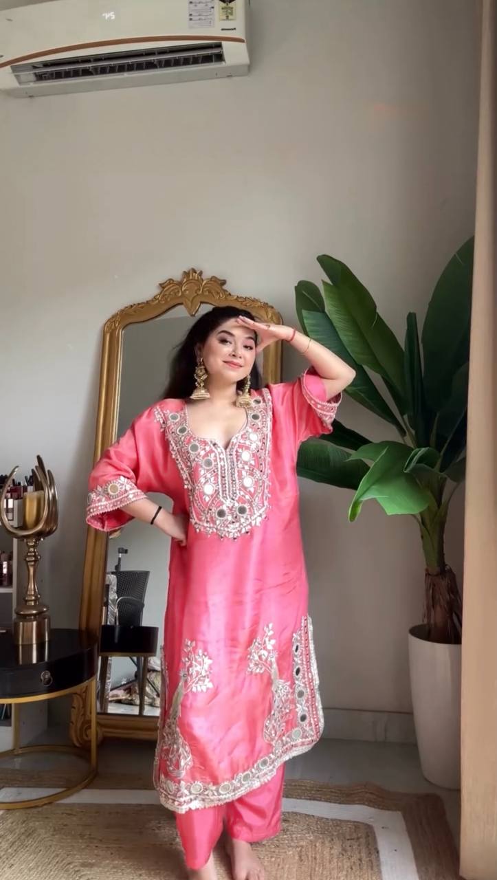 Woman in a pink traditional outfit standing in a room with a mirror and plant.